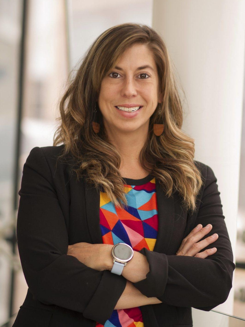 A woman with brown hair and a black blazer and colorful blouse stands in a doorway with her arms folded, smiling